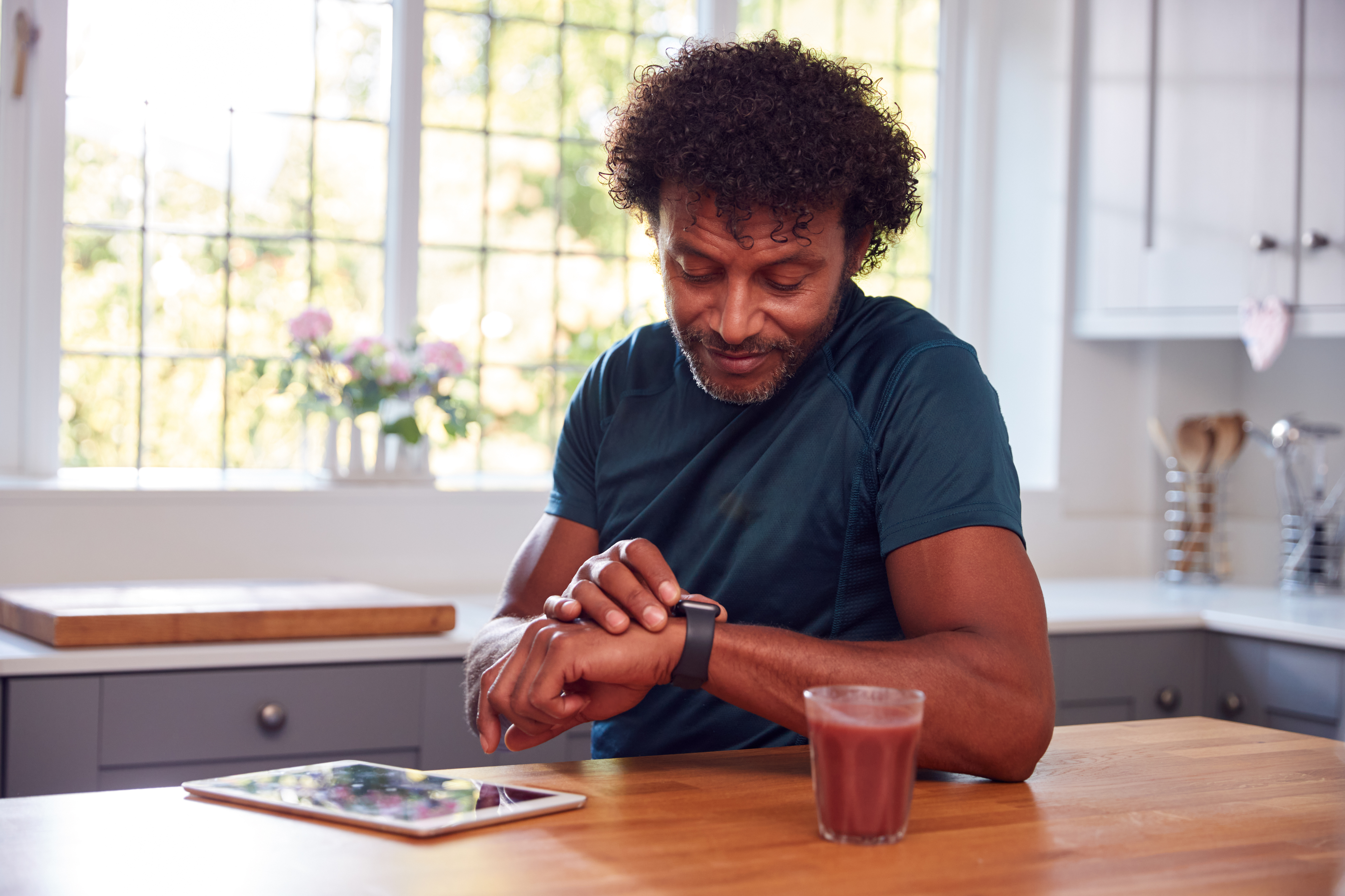 Mature Man Wearing Fitness Clothing At Home Logging Activity From Smart Watch Onto Digital Tablet