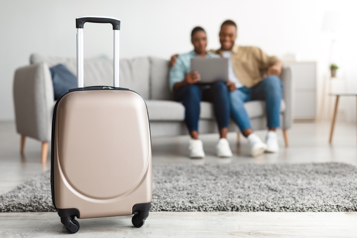 Tourists Couple Using Laptop Booking Tickets Online Sitting On Couch At Home. Selective Focus On Luggage