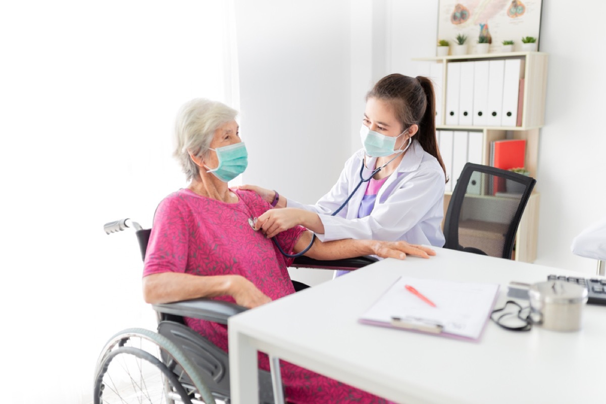 young female doctor with face mask checking the heartbeat of older woman with face mask in wheelchair