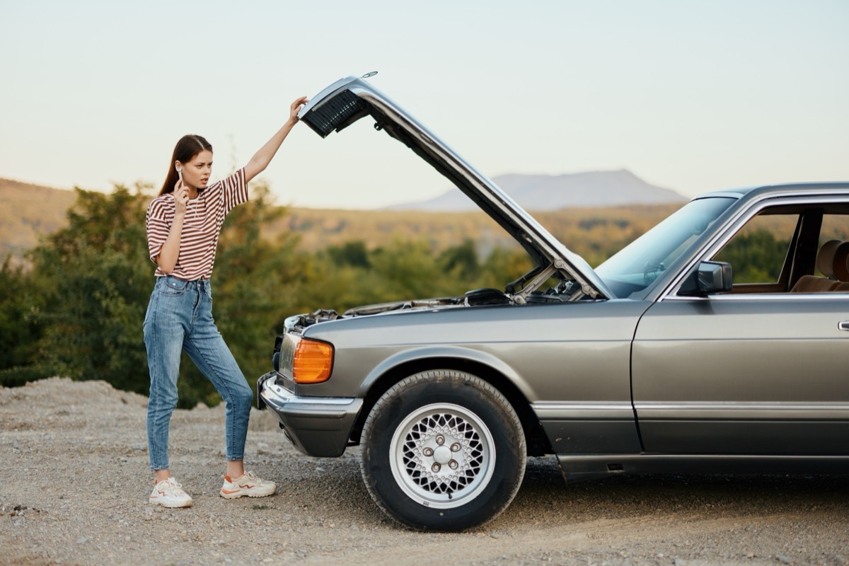 woman standing on the side of the road next to her old, broken-down car and calling for help