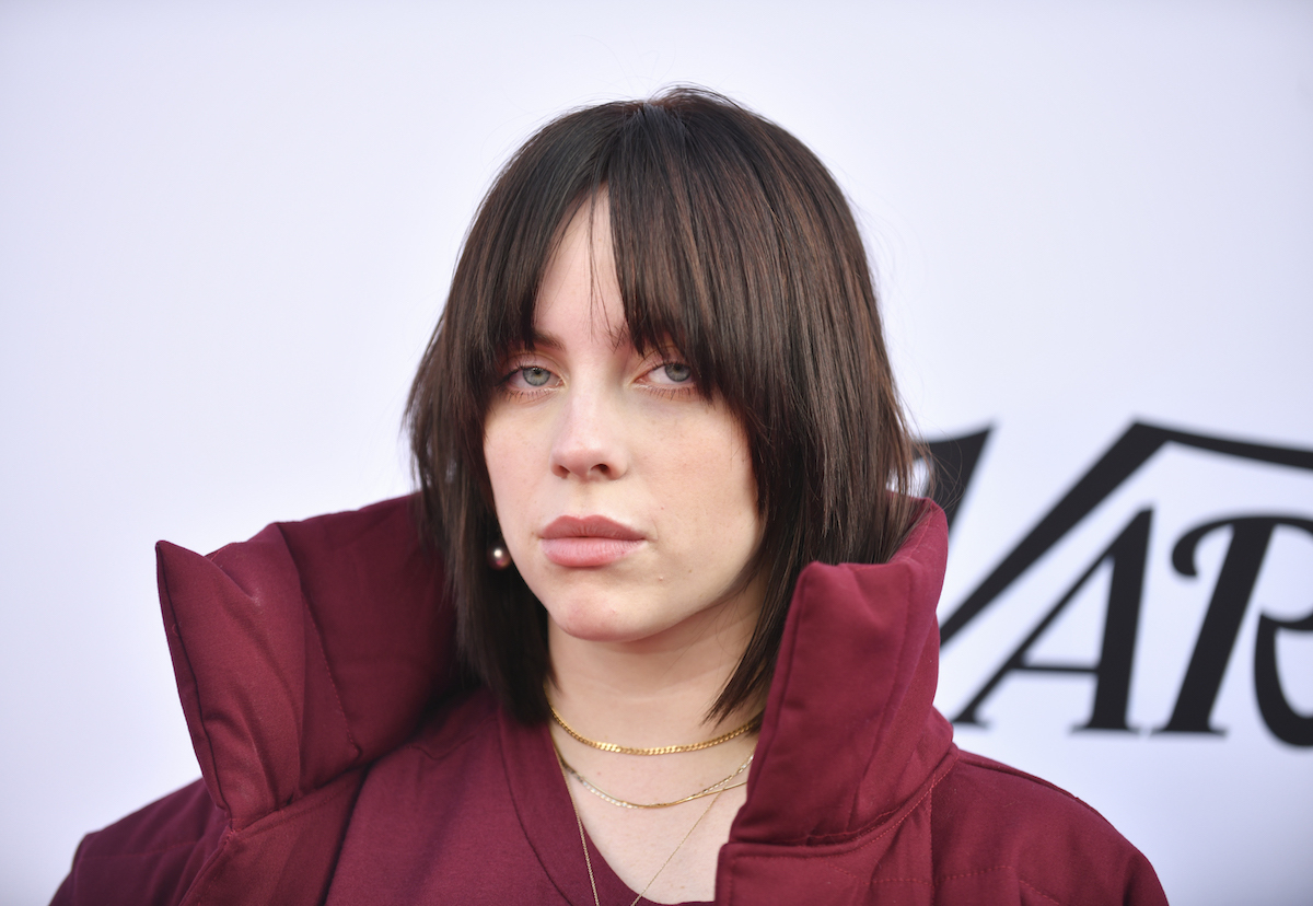 Billie Eilish posing at a Variety event wearing a red coat with a large collar; her brunette hair is styled in a wolf cut