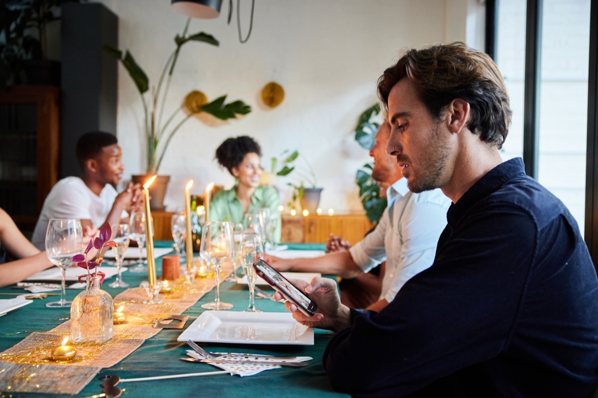 Young man distracted by a text on his mobile phone while sitting at a table with friends before an evening dinner party