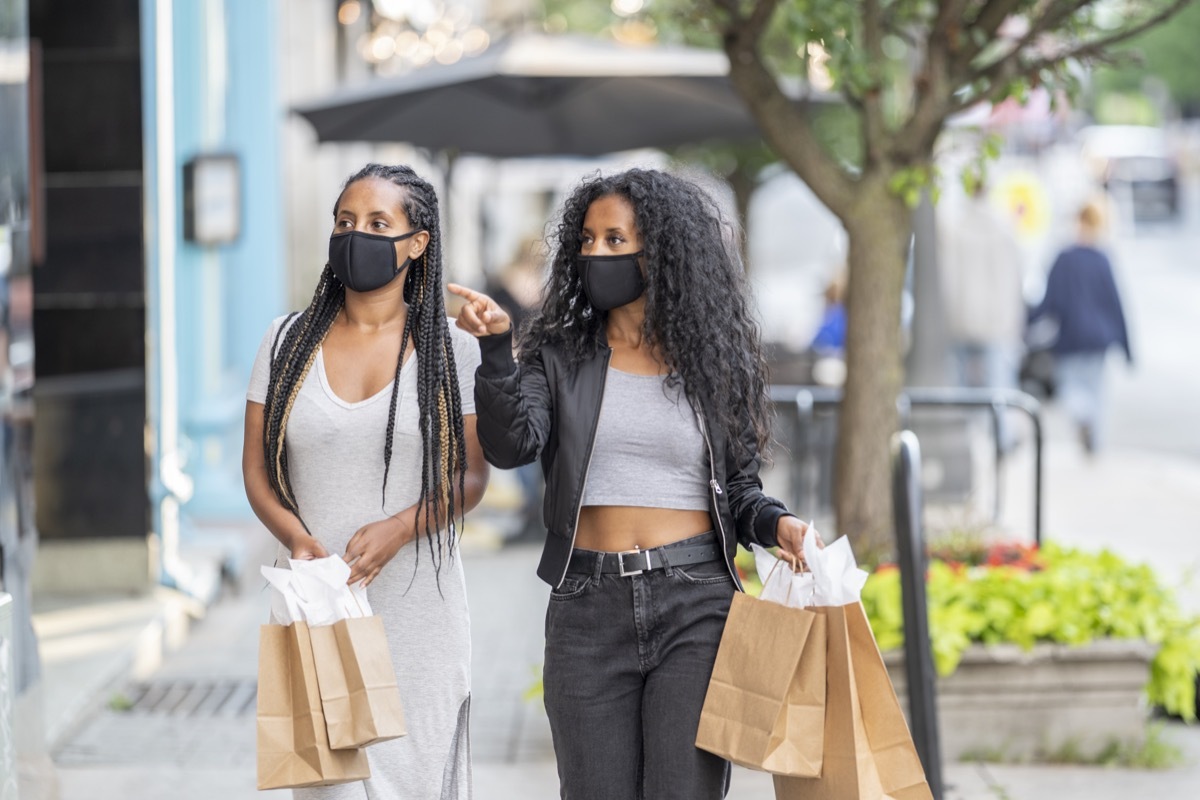 Two woman walking downtown, holding shopping bags, and doing some window shopping while wearing protective face masks during the COVID-19 pandemic.