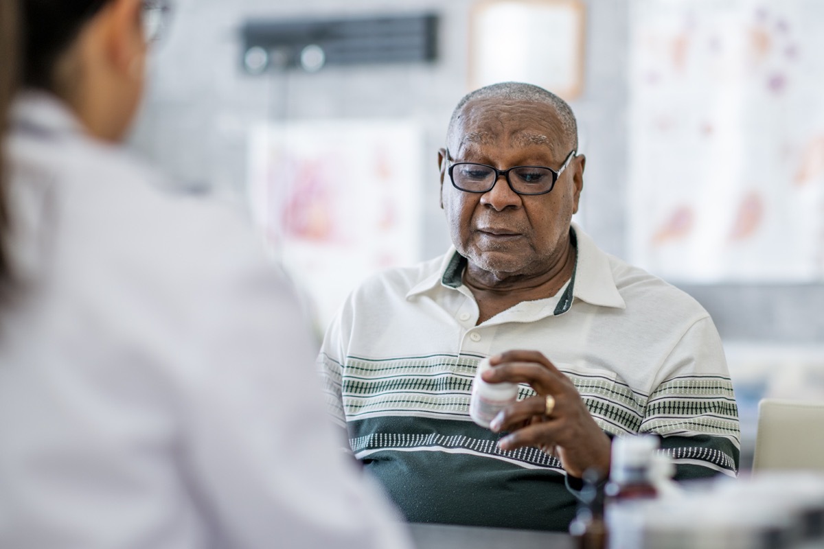 An elderly black man looks at a pill bottle in his hand while sitting across the table from a doctor in the doctor's office. He appears to be making a decision about it.